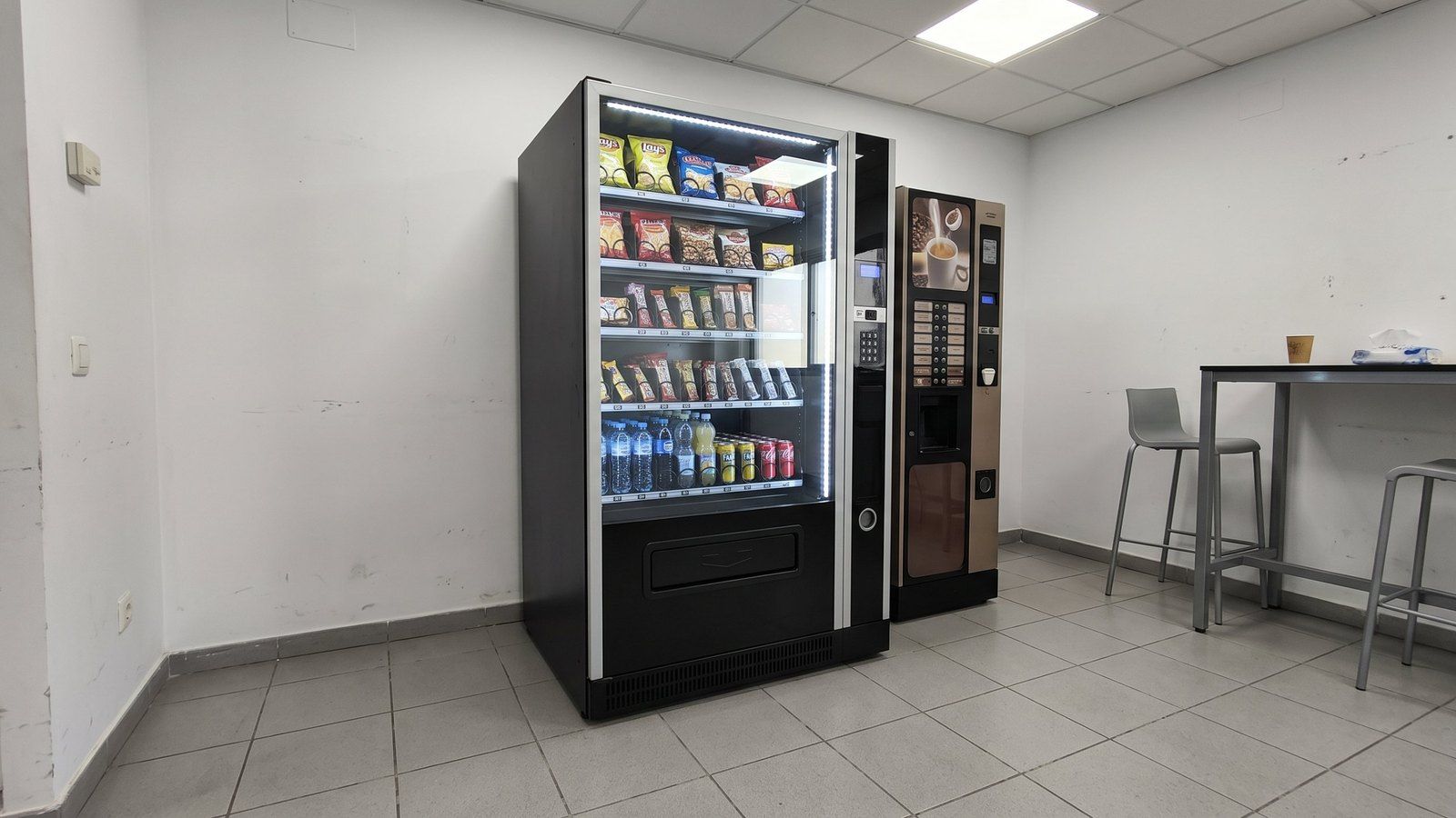 Máquina de vending mixta para empresas con snacks y bebidas frías junto a una máquina de café en la sala de descanso de una oficina